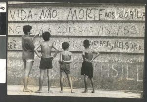 Quatro crianças descalças, três deles sem camisa, observando um muro pichado durante o período de ditadura no Brasil.